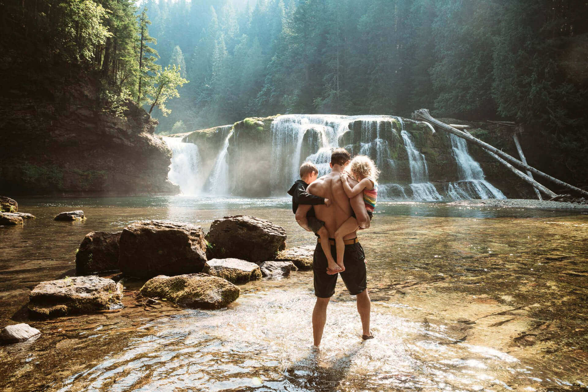 A man holders two small children while standing in the water in front of Lower Lewis Falls.