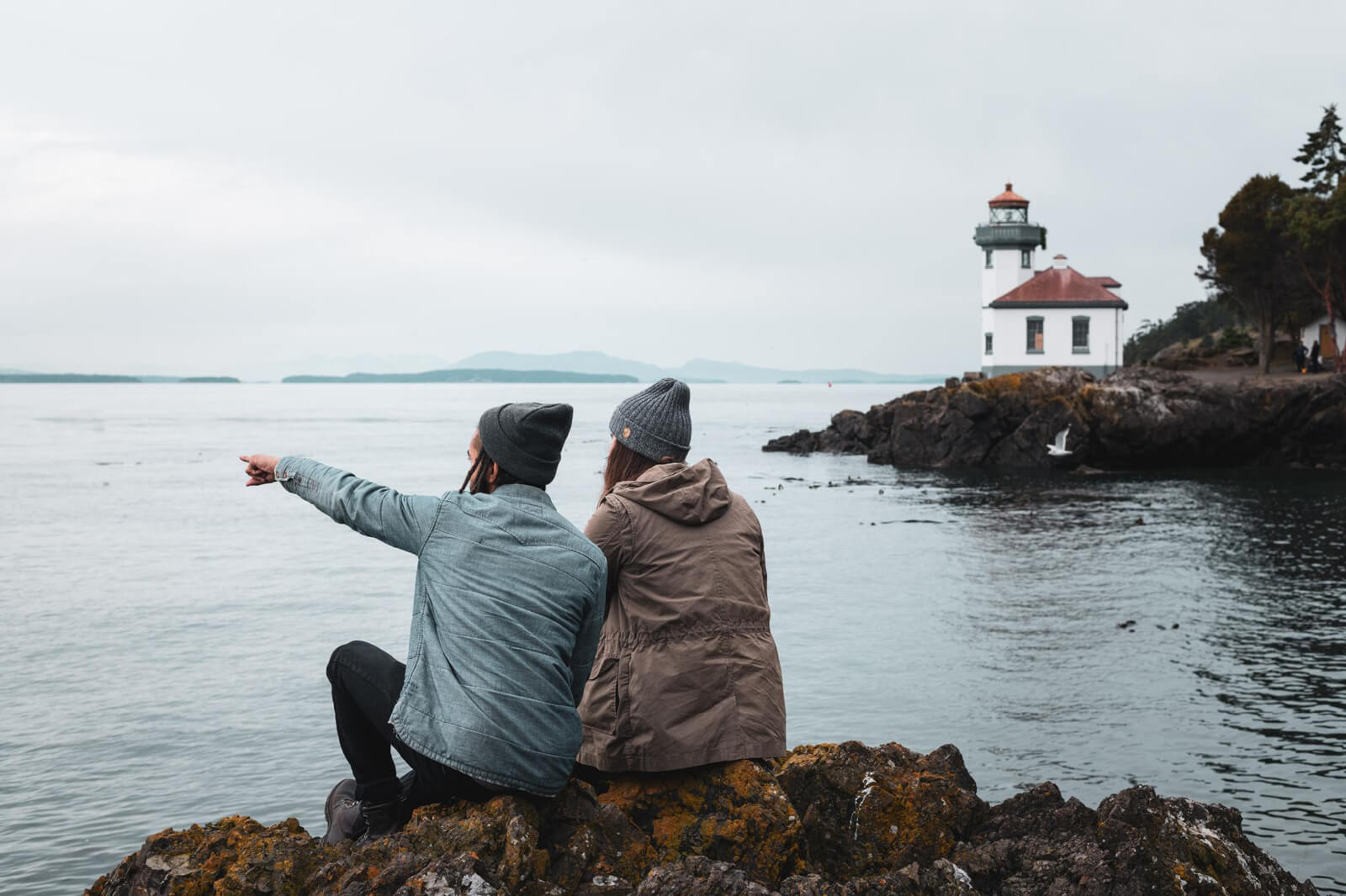 A couple sits on rocks near a lighthouse on San Juan island, a romantic spot in Washington