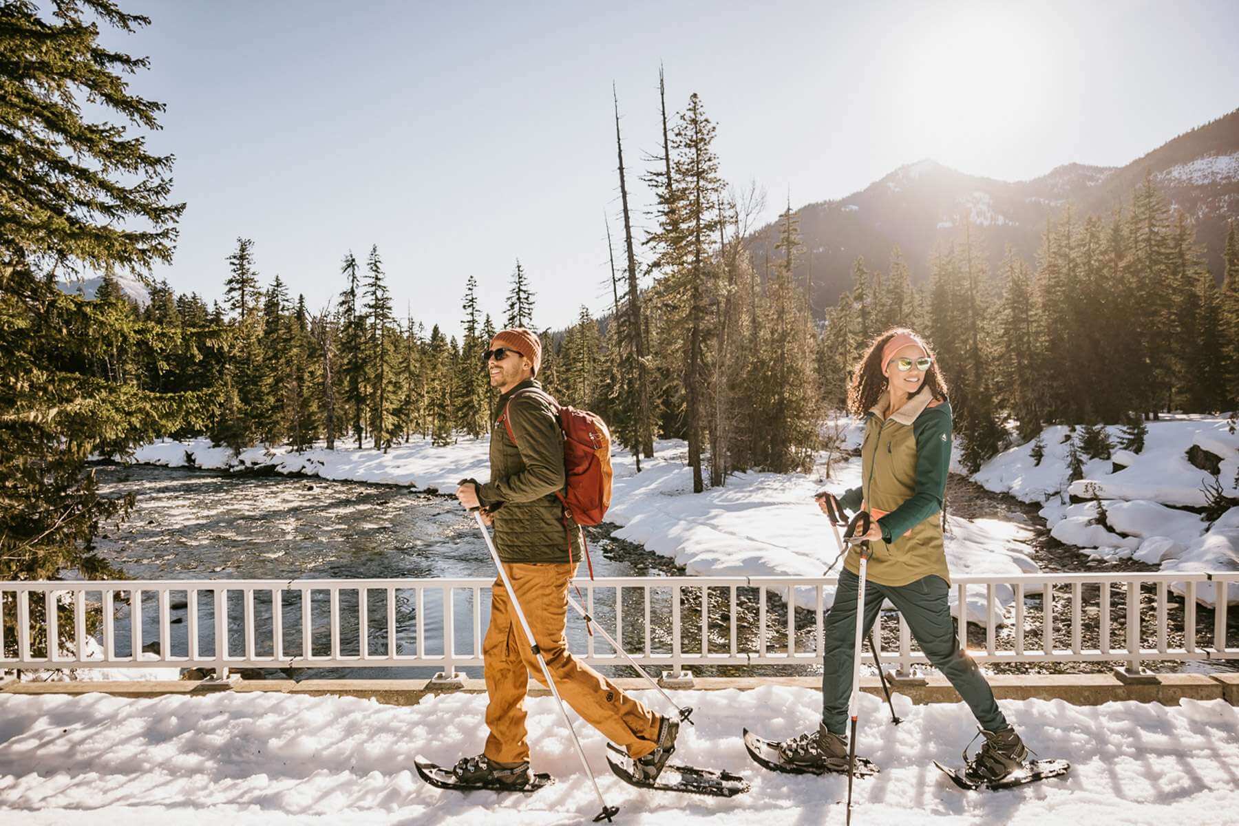 A man and woman snowshoe across a snow-covered bridge over a river during a winter getaway in the Cascade Mountains