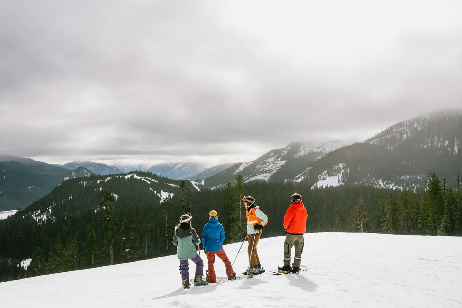 Four people in ski gear stand in the snow looking down from a mountain