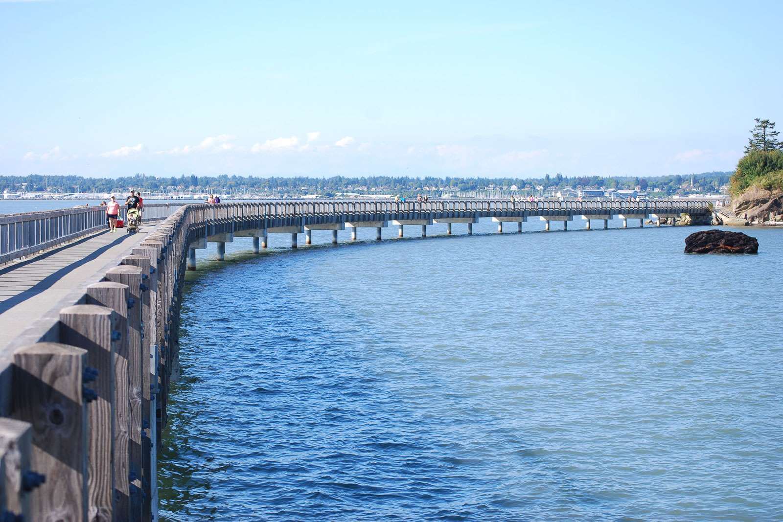 People walk along a wooden and concrete boardwalk that extends over the water in Bellingham