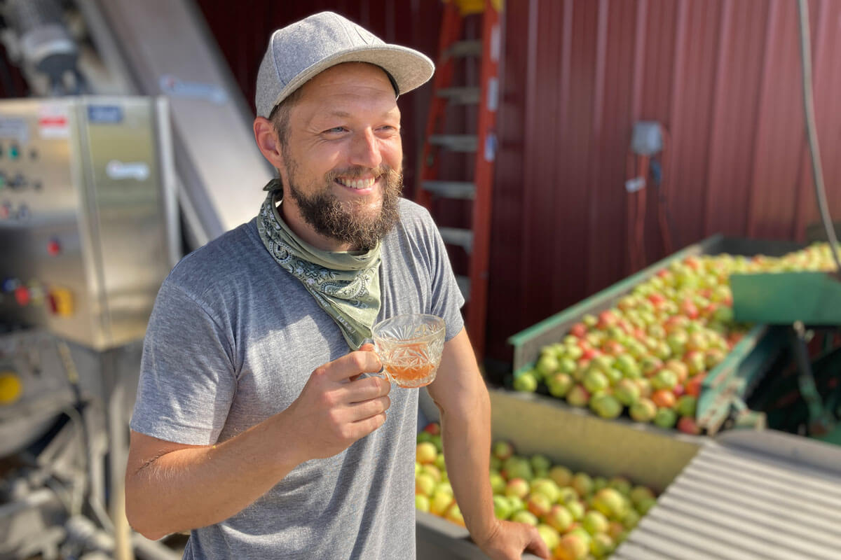 A man holds a glass of fresh-pressed cider in front of a bin of apples at Finnriver Farm & Cidery