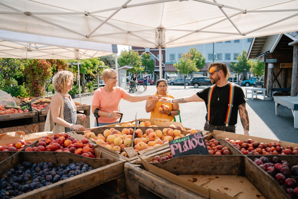 A mean wearing rainbow suspenders over a black shirt hands out sliced peaches to a group of three women at the Olympia Farmers Market. 