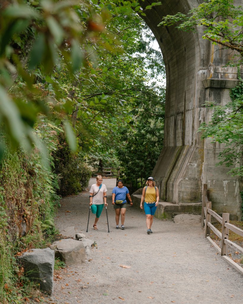 Three women walk under a bridge overpass along a tree-lined trail. 