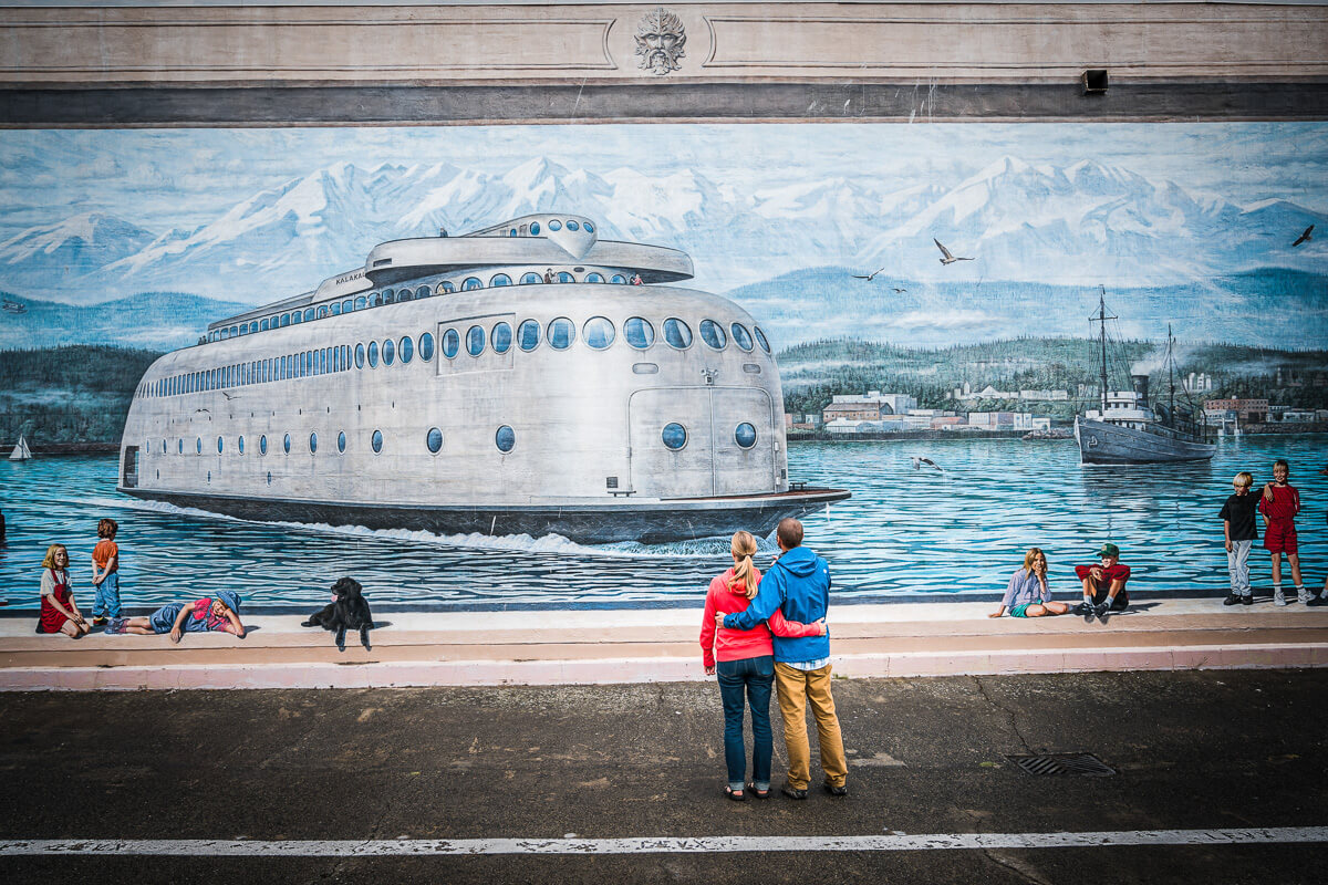 A couple stands arm in arm in front of a mural