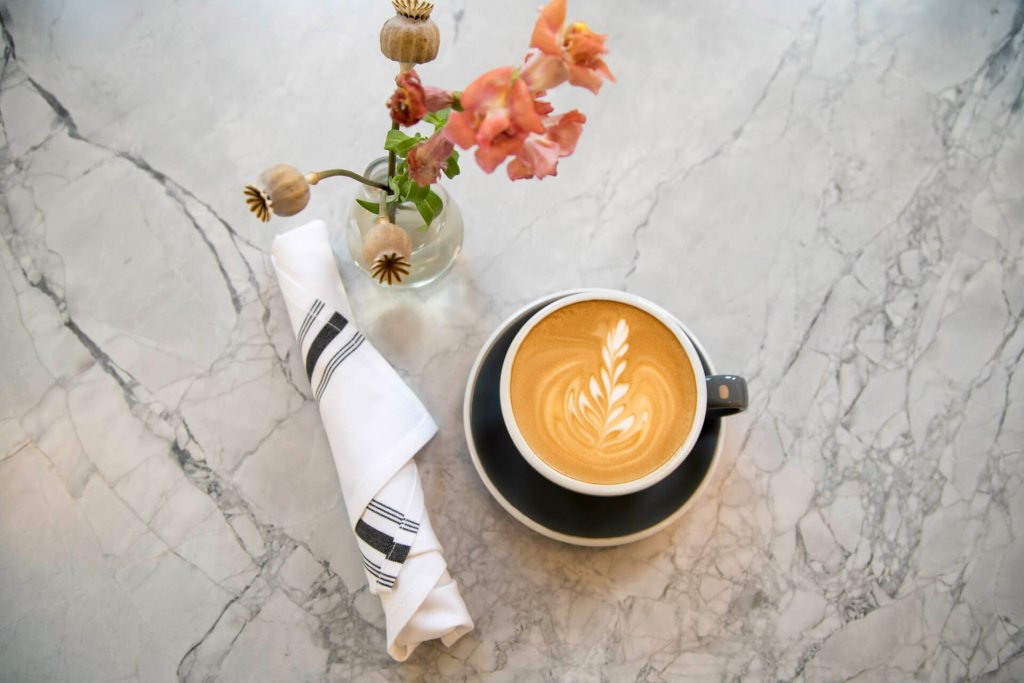 Washington State Coffee Roasters: A rolled napkin, latte, and a glass vase with flowers sit on a marble table at Camber Coffee