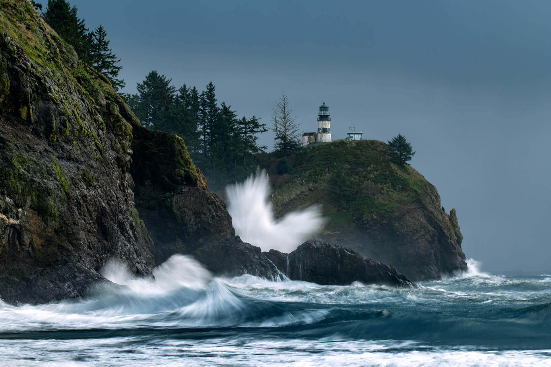 Cape Disappointment Lighthouse stands on a rocky cliff as waves crash against the rocks.