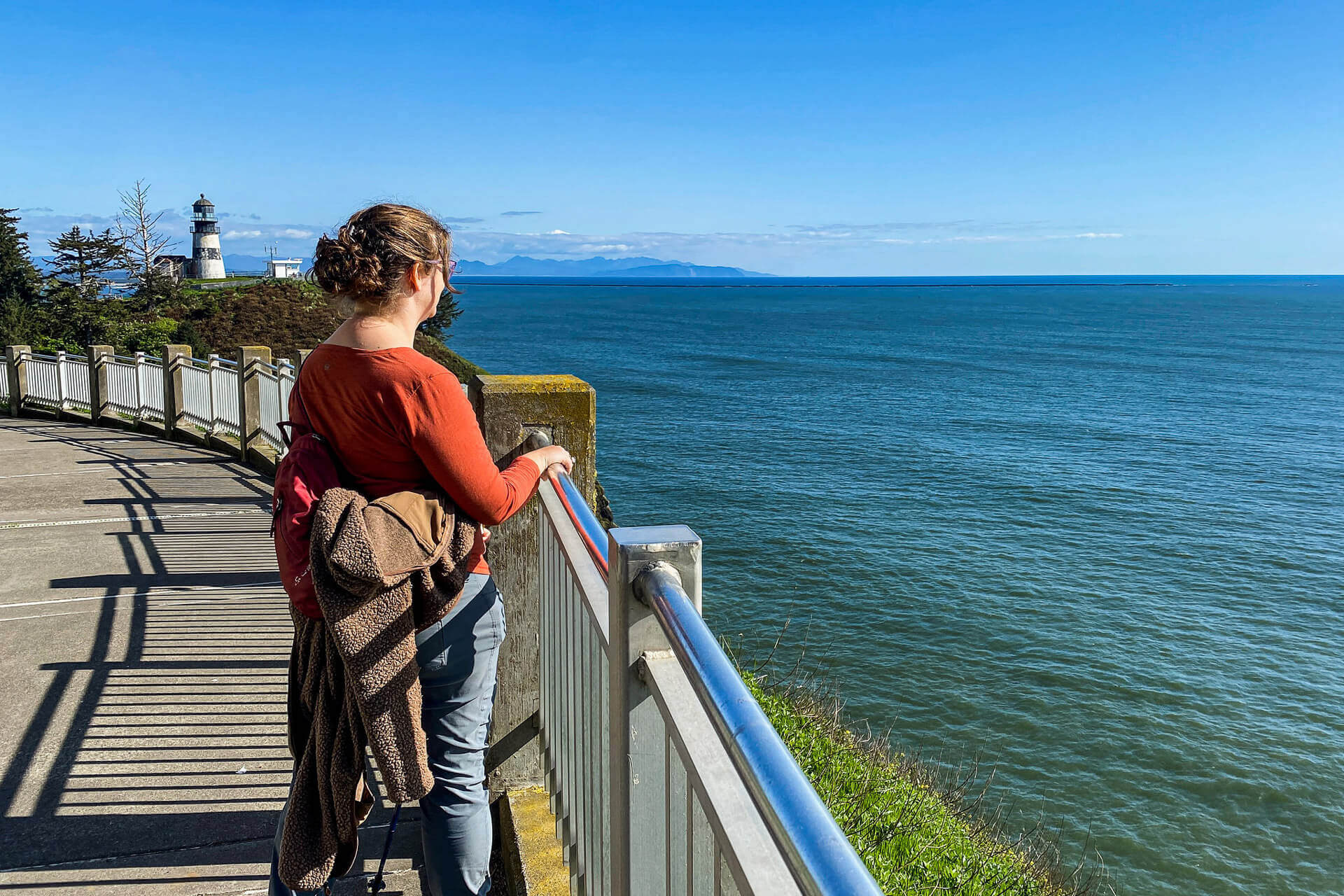 A woman stands behind a railing overlooking the water with a lighthouse in the background