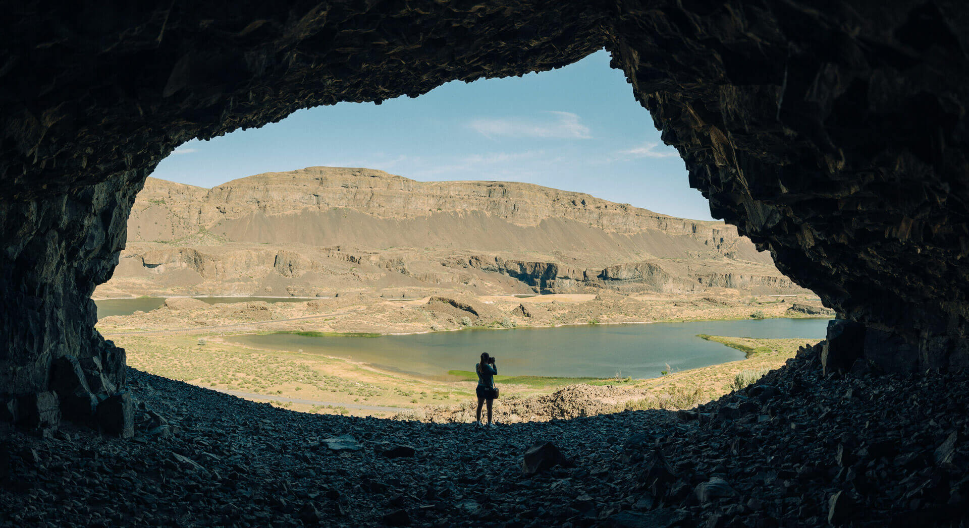 A stands in the entrance to a cave and takes a photo with brown canyons in the background. 