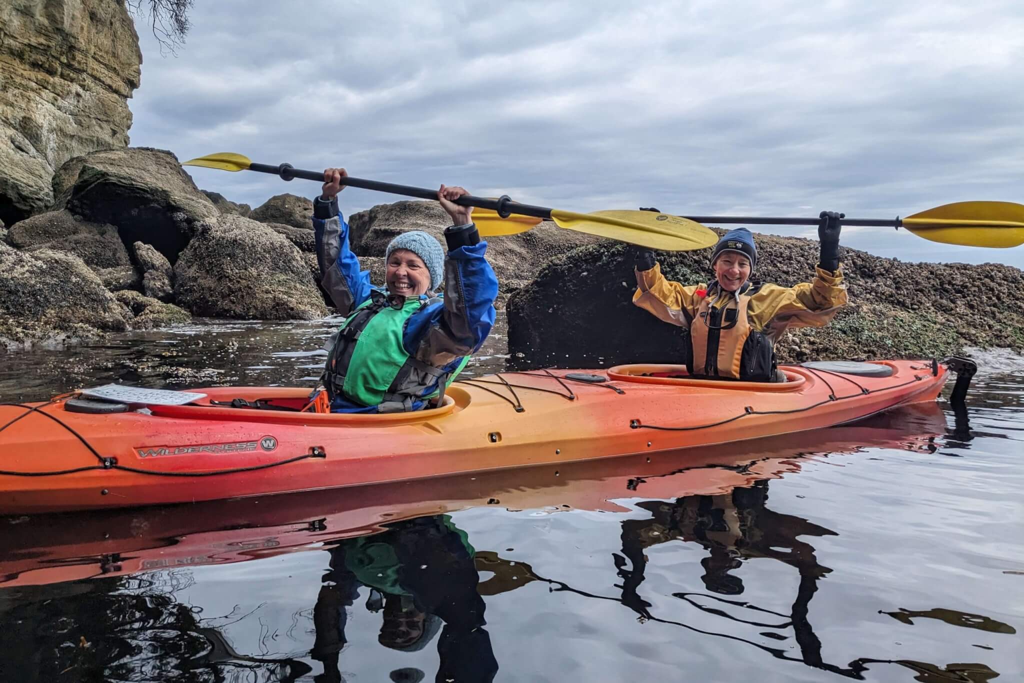 Two people wearing long-sleeved coats, lifejackets, and beanies raise their paddles above their heads near a rocky shore. 