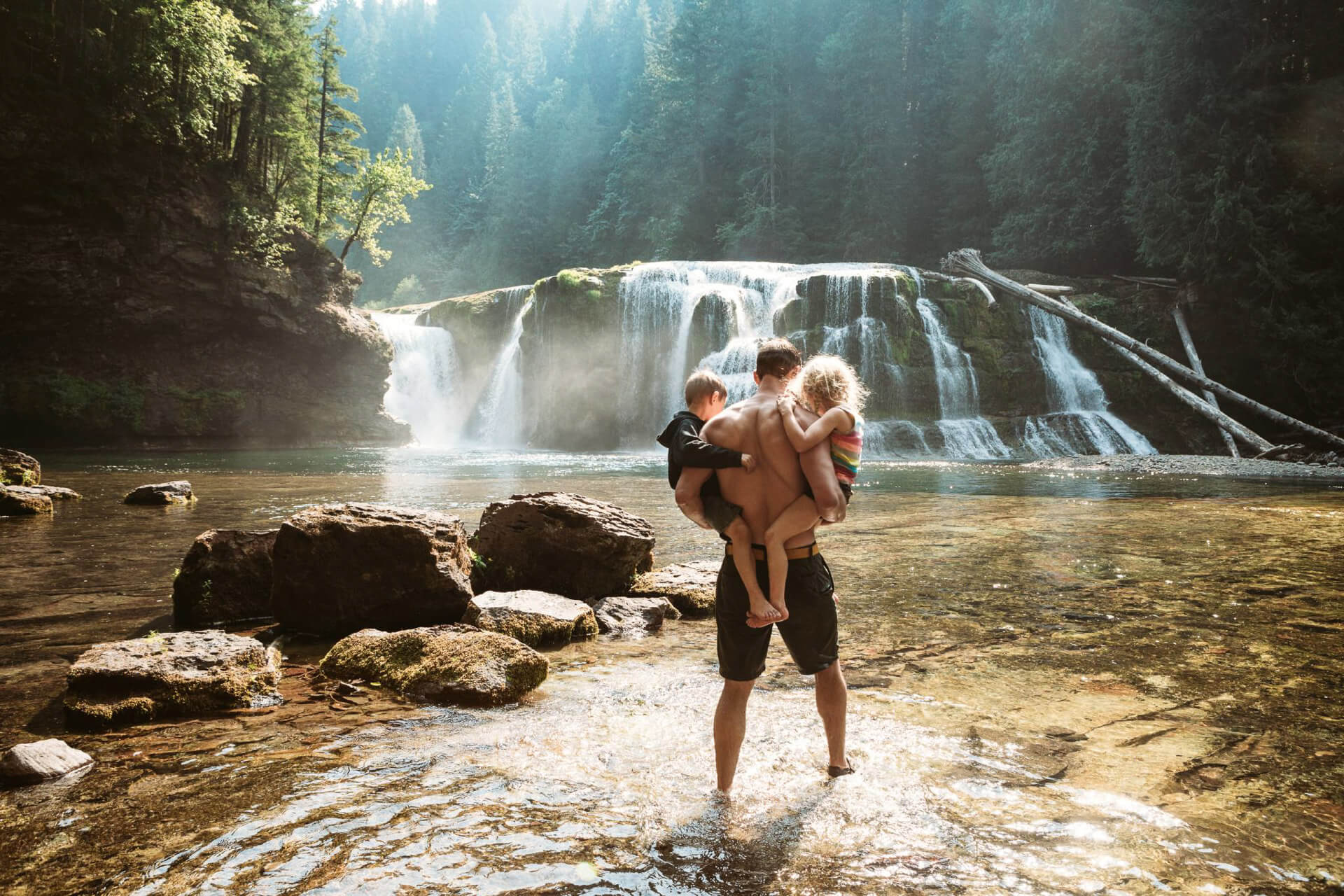 A man wades in front of a waterfall as he holds two children at Lower Lewis Falls, a swimming hole in Washington.