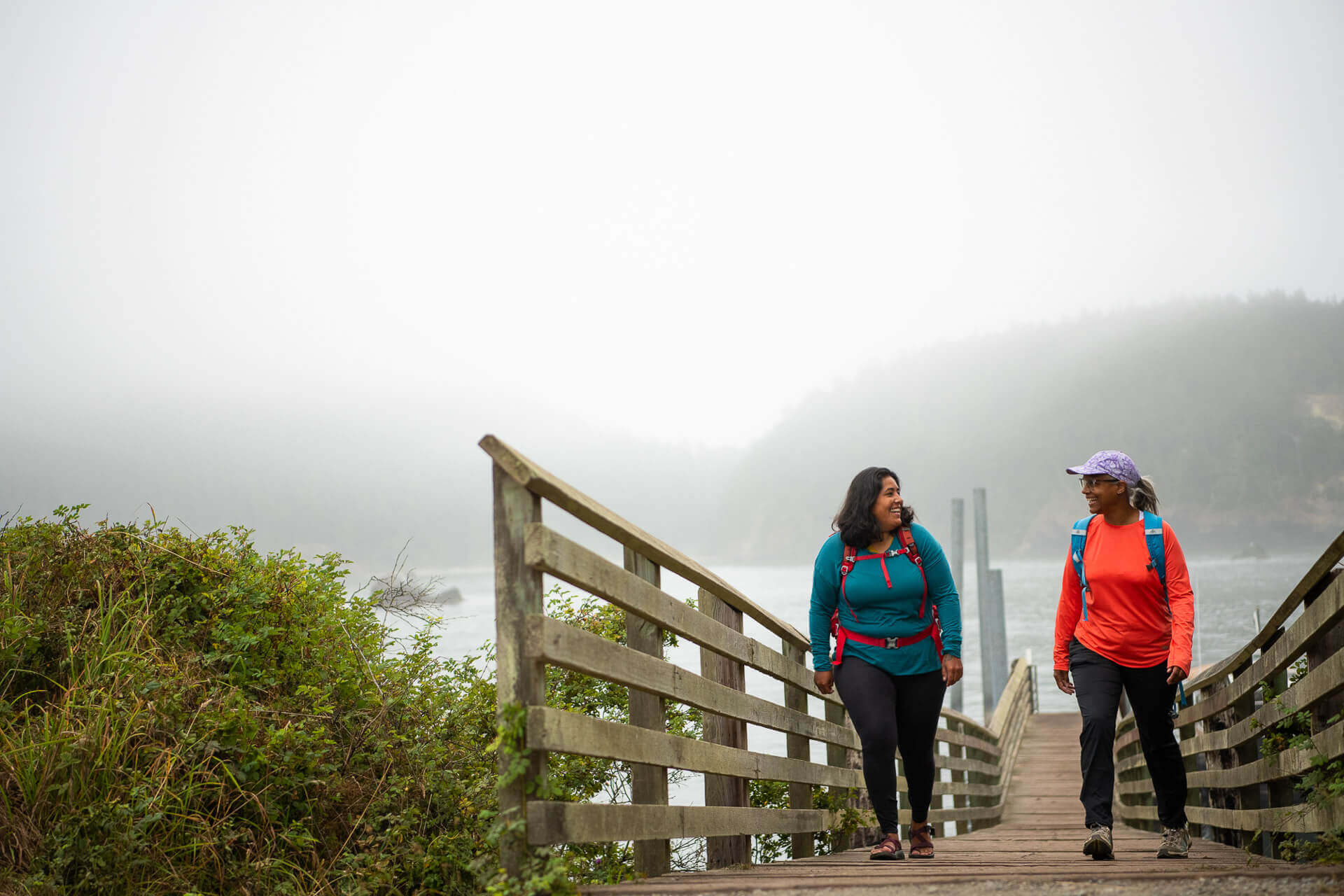 Two women wearing backpacks walk up a dock in the fog at Deception Pass State Park.