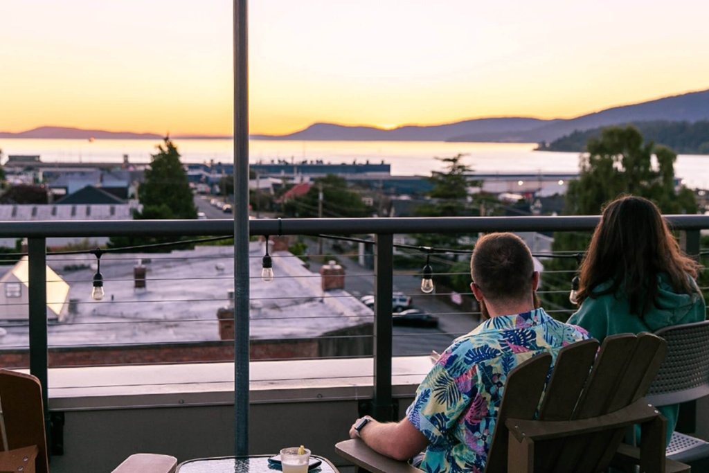 A man sits on the rooftop deck of the Majestic Inn overlooking the water at sunset.