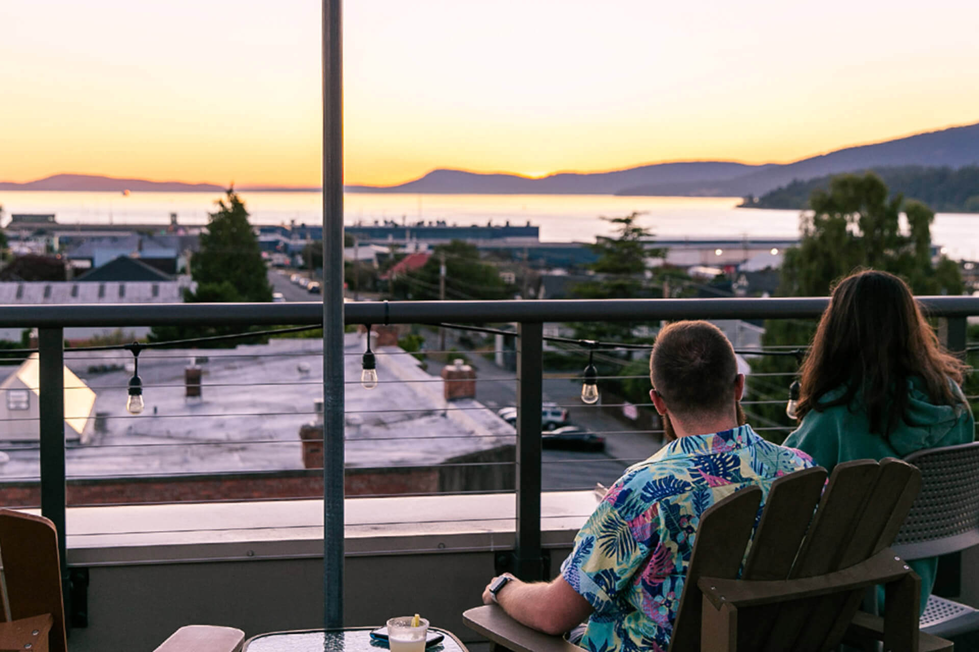 A man and woman sit on the rooftop deck of the Majestic Inn & Spa overlooking the water at sunset.