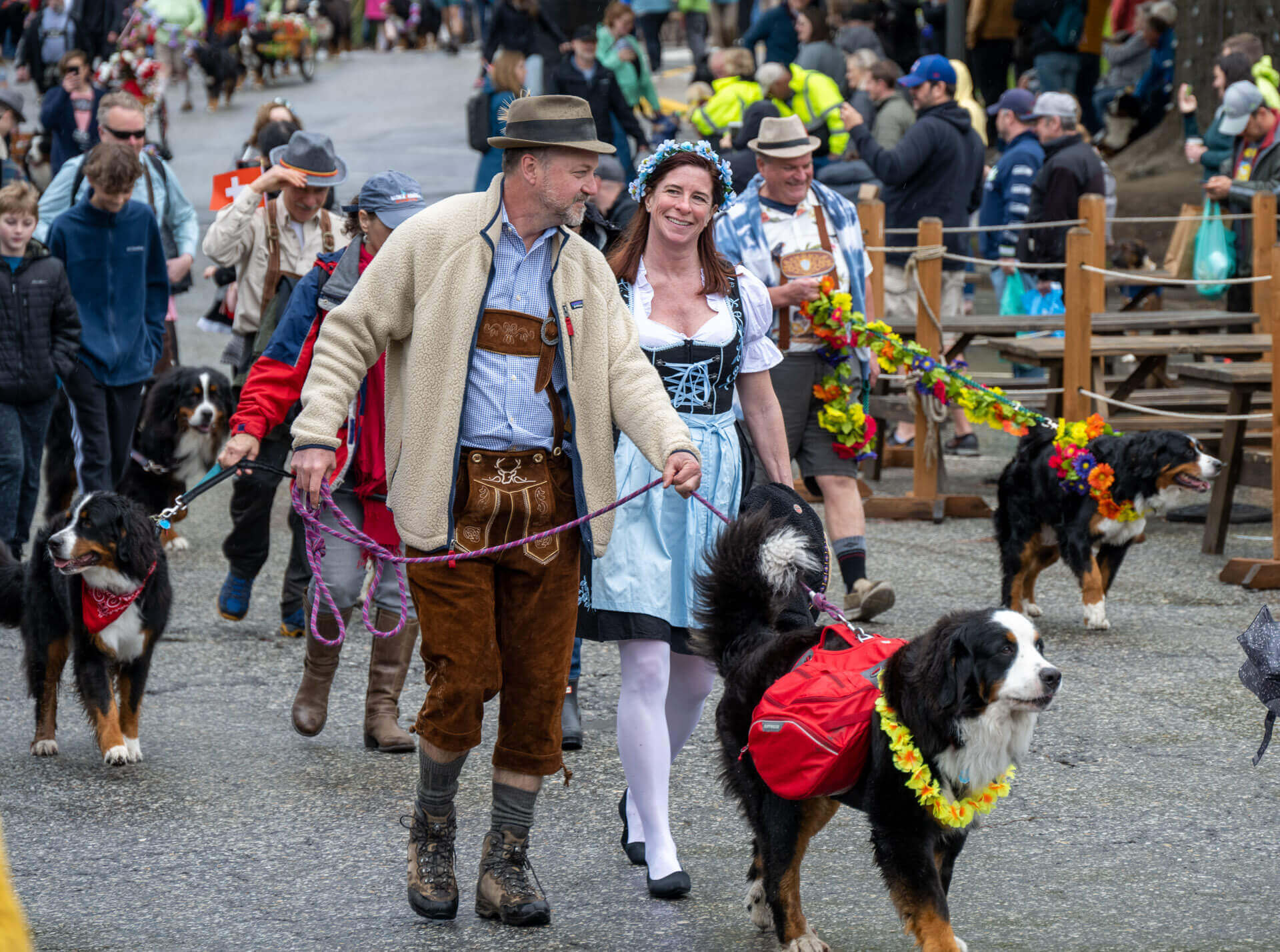 A man and woman wearing traditional German outfits walk a dog wearing a flower necklace and red backpack in Leavenworth Washington