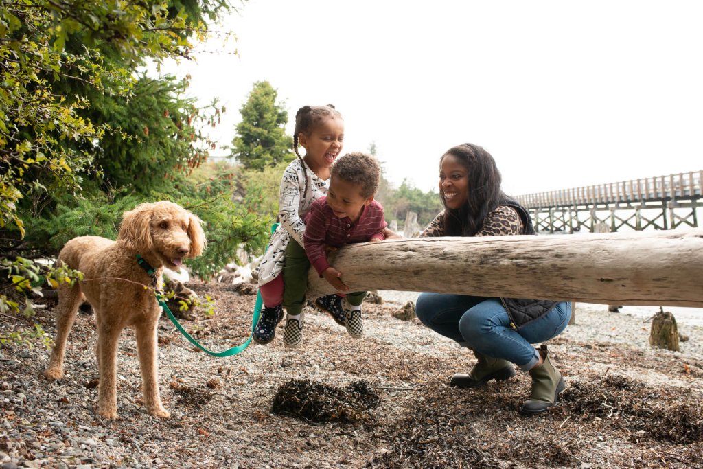 Two children play on a log on a beach in Bellingham while a mom and dog watch them
