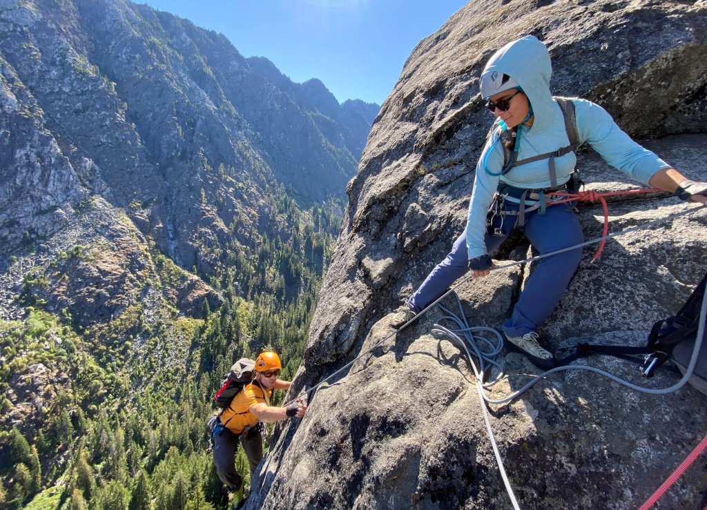Two people in helmets and gear use a rope to climb a rock cliff.