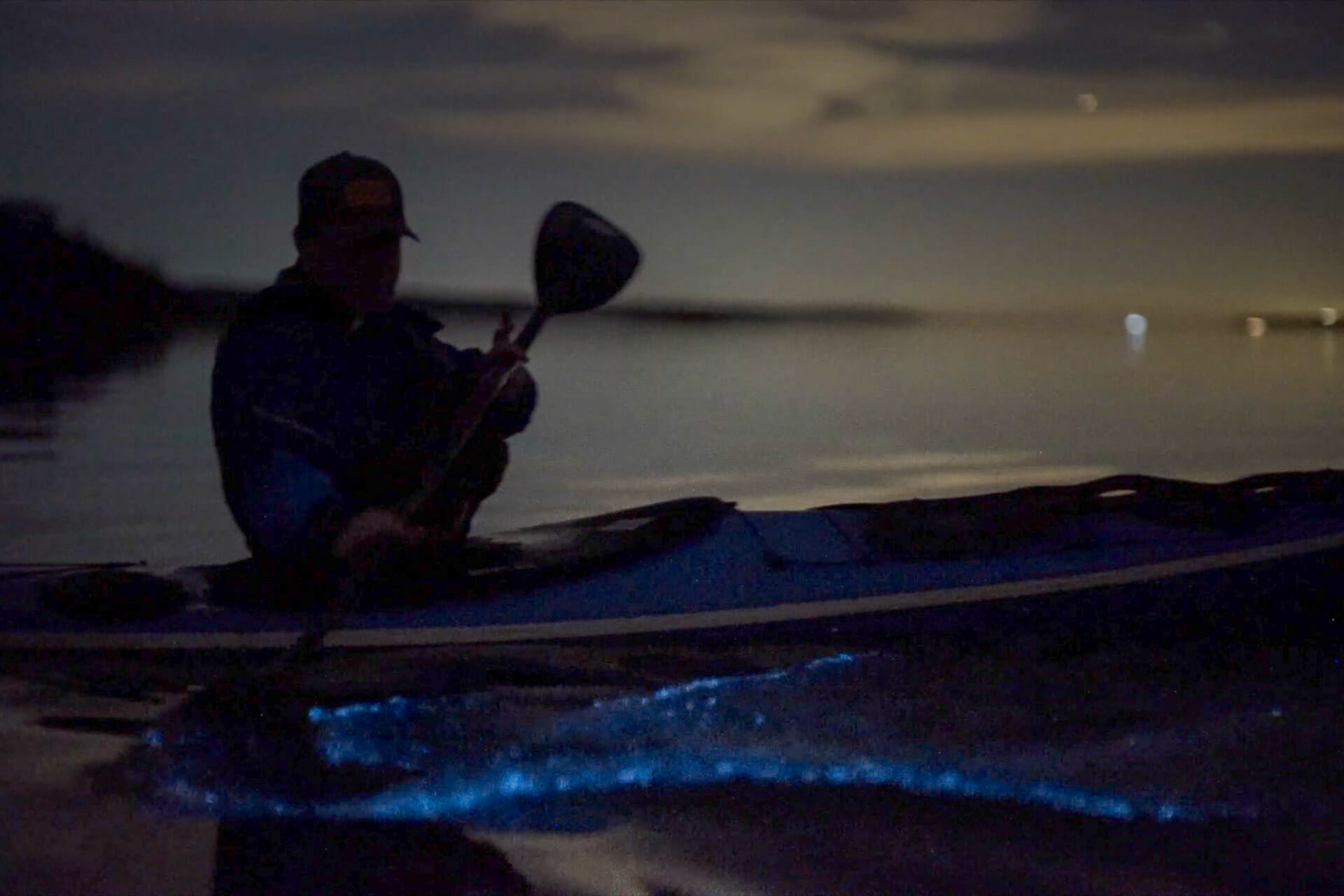 A kayaker disturbs water with his paddle at night, causing bioluminescence.