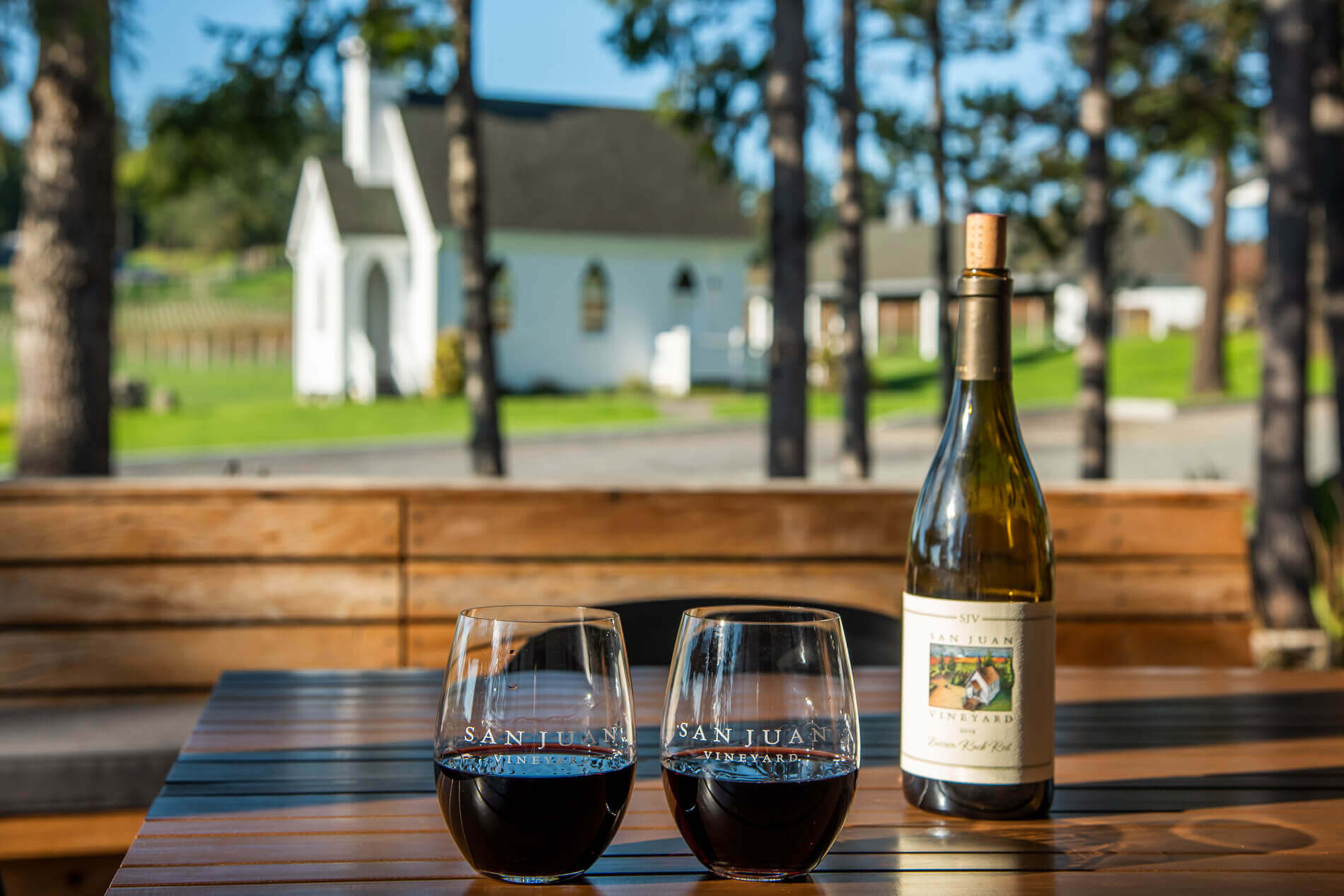 A photo of two wine glasses filled with red wine sitting on a wooden table outside at San Juan Vineyard.