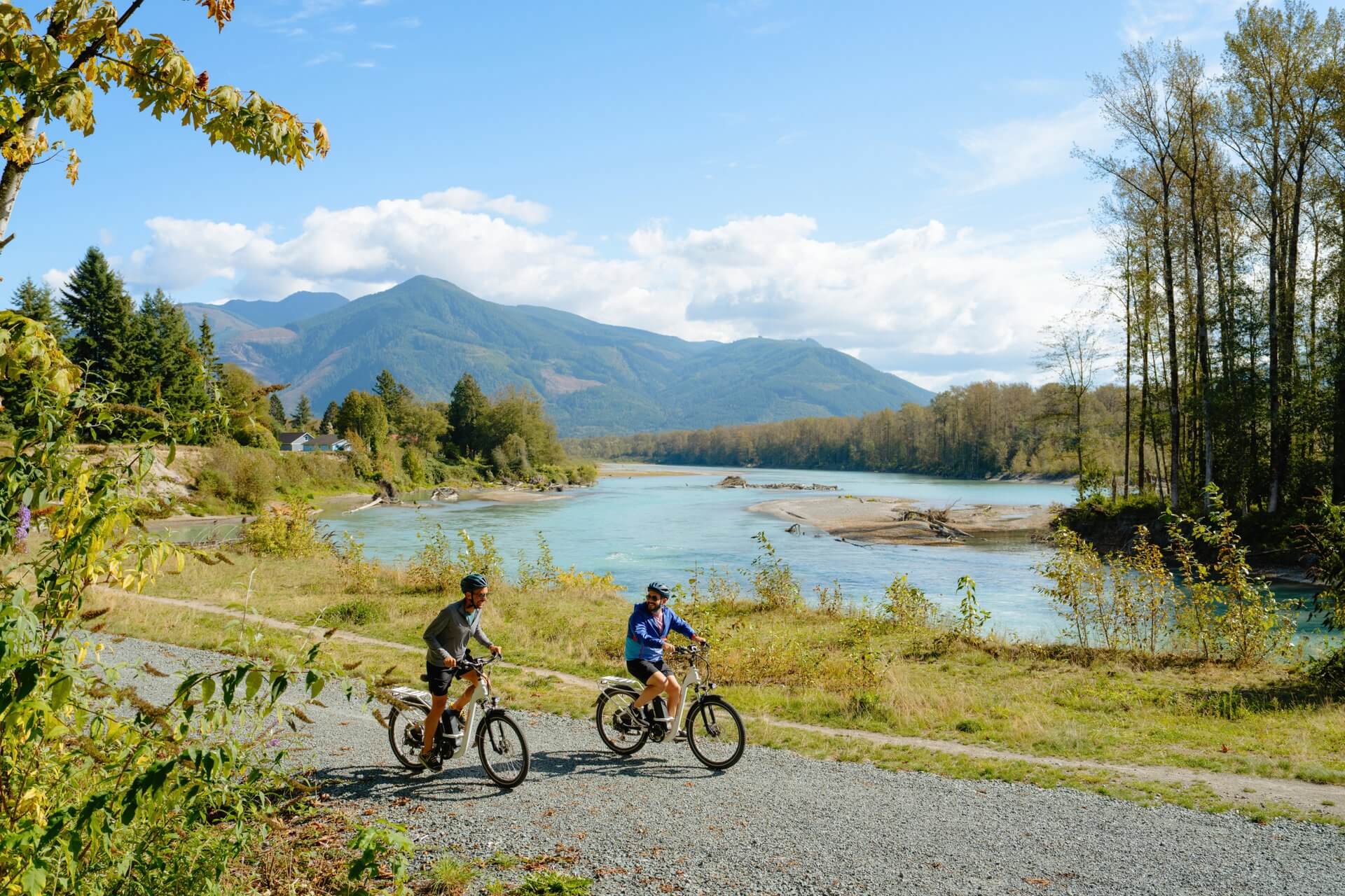 Two men bike on a gravel path along the Skagit River with mountain foothills in the background.