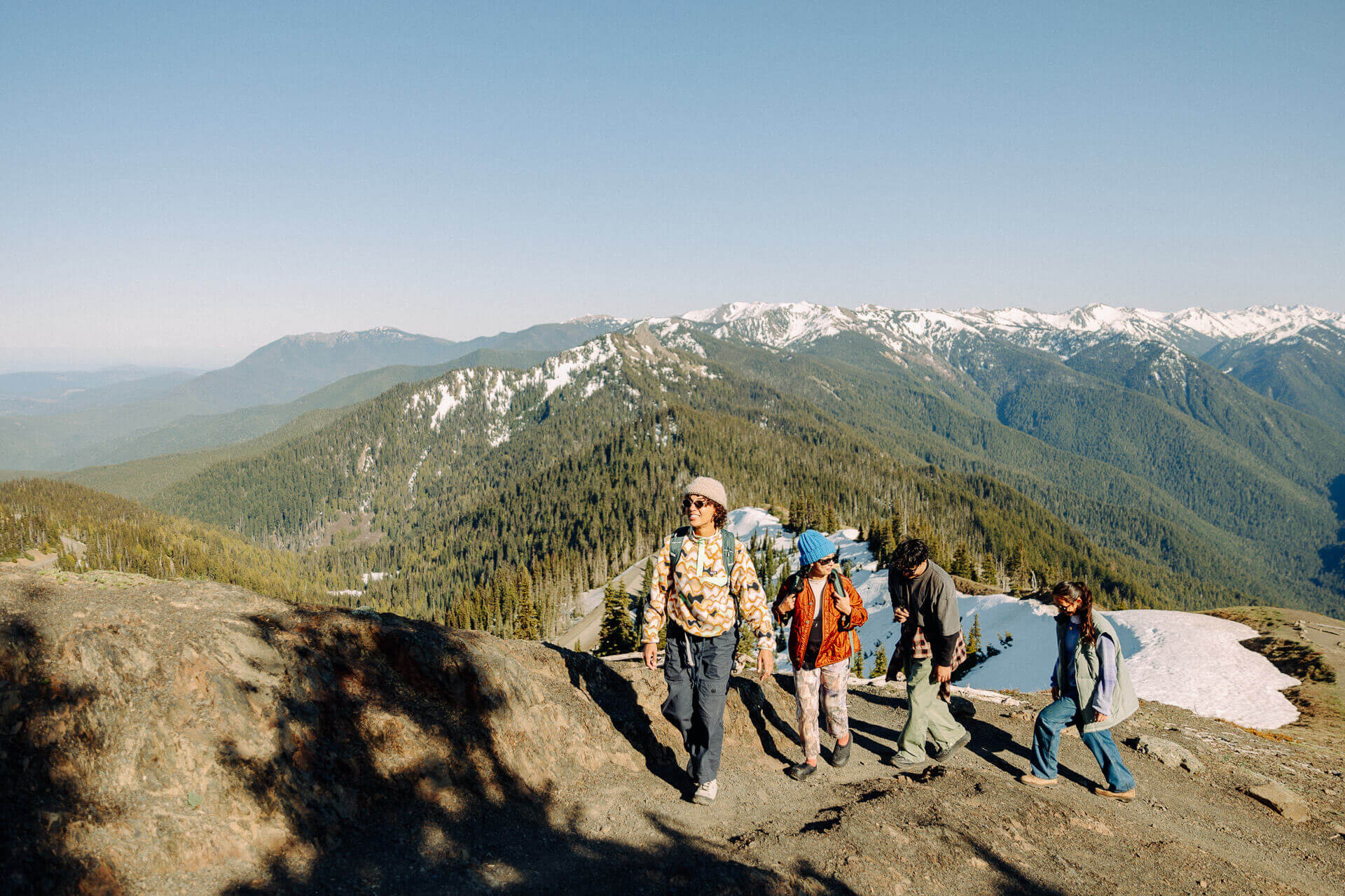A group of four people hike along a trail with snow-dusted mountains in the background