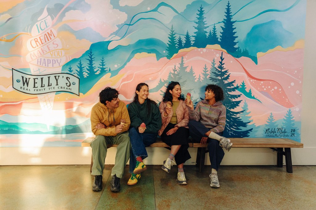 A group of four people sit on a bench and eat ice cream in front of a mural