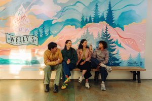 A group of four people sit on a bench and eat ice cream in front of a mural