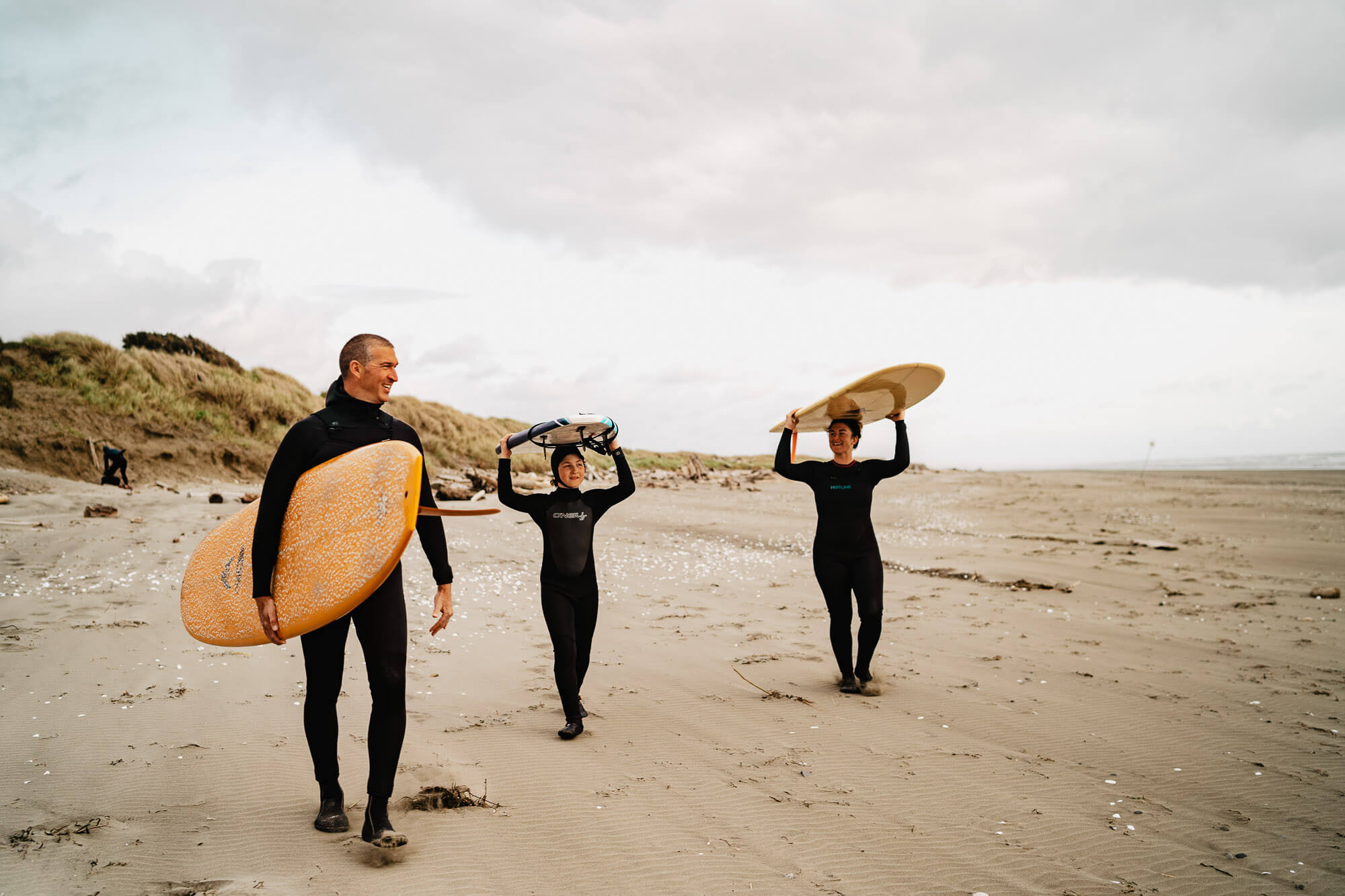 Three people in wet suits carry surfboards on the beach in Westport